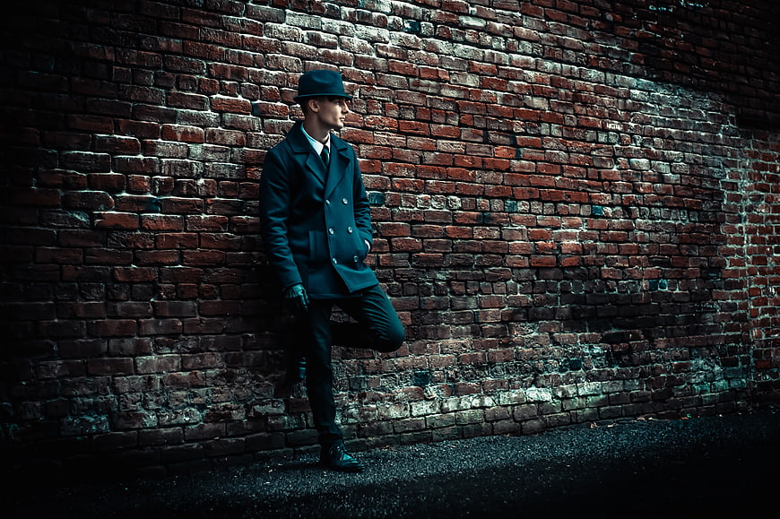 Stylish man in hat and coat leaning against brick wall