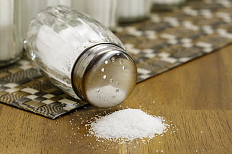 Tilted glass salt shaker with spilled salt on wooden table