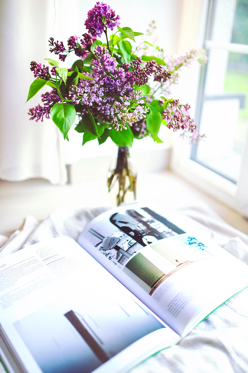 Purple lilac flowers in vase with open book on bed