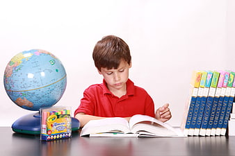 Young boy reading book at desk with globe and school supplies