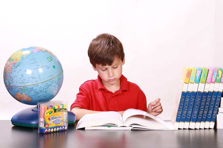 Young boy reading book at desk with globe and school supplies