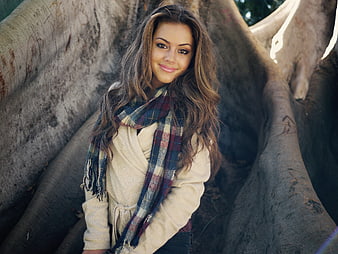 Smiling young woman with plaid scarf and beige sweater leaning against tree