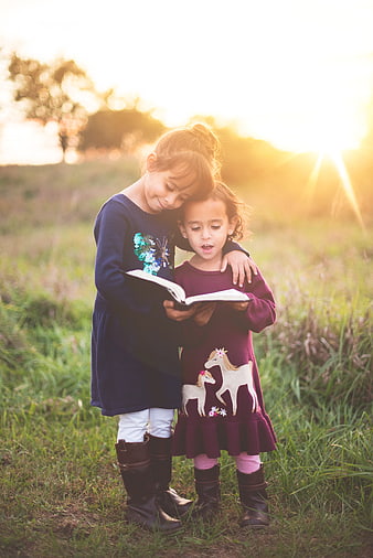 Two young girls reading a book together in a grassy field at sunset