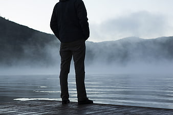 Man standing on wooden dock overlooking misty lake and mountains