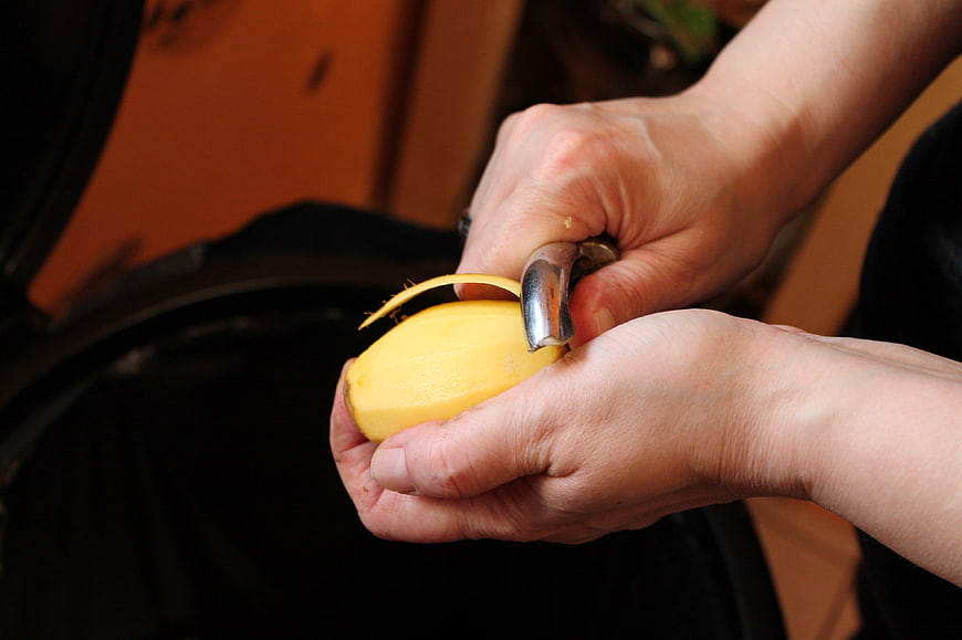 Hands Peeling A Yellow Potato With A Metal Peeler In Kitchen
