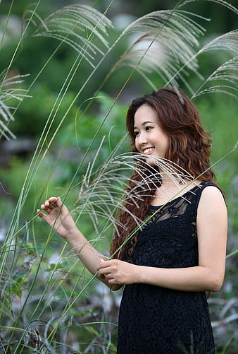 Smiling young Asian woman in black dress touching tall grass outdoors