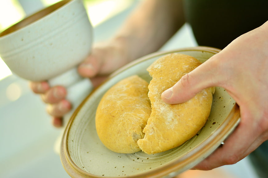 Hands breaking bread on ceramic plate with cup in background
