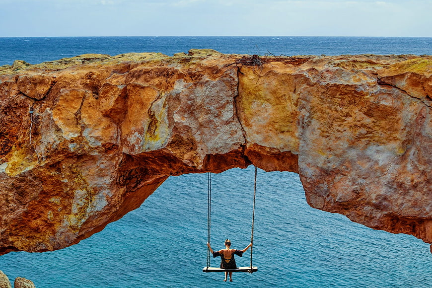 Woman on swing under natural rock arch overlooking ocean