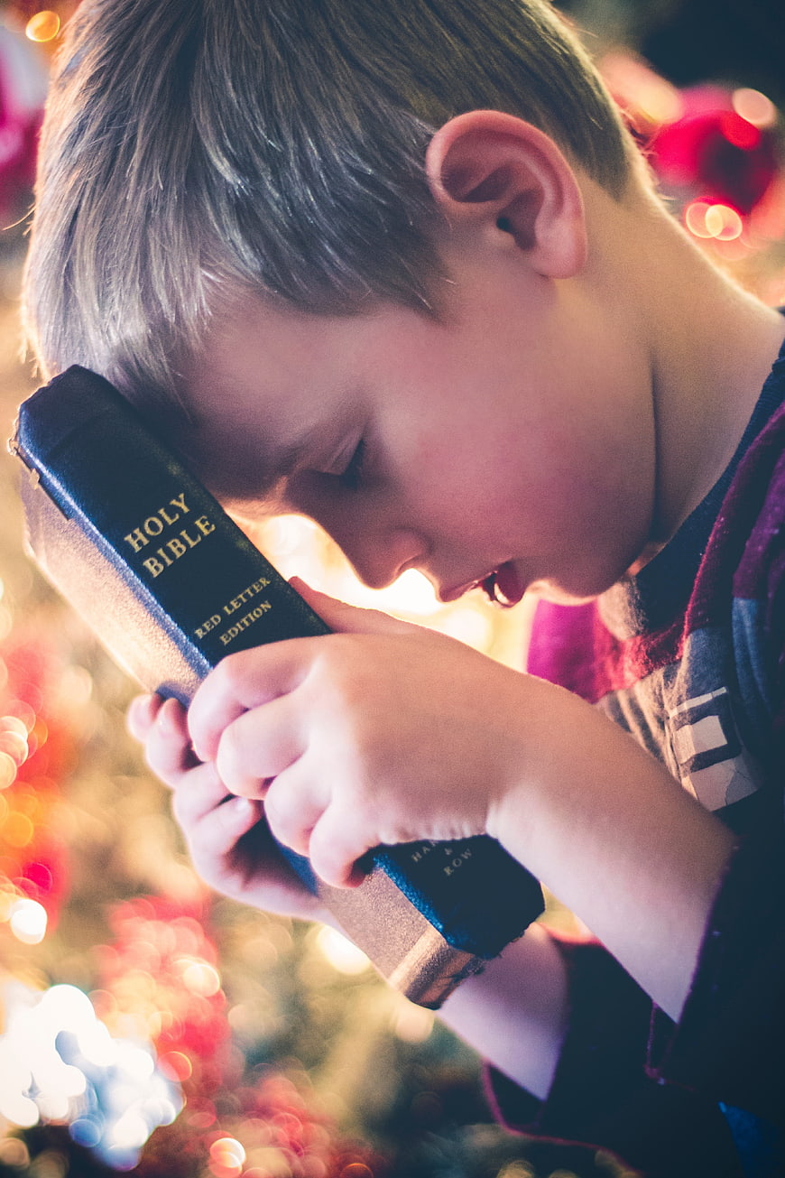 Young boy praying with Holy Bible near Christmas tree