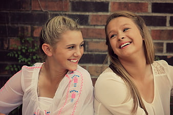 Two smiling teenage girls in white tops against a brick wall