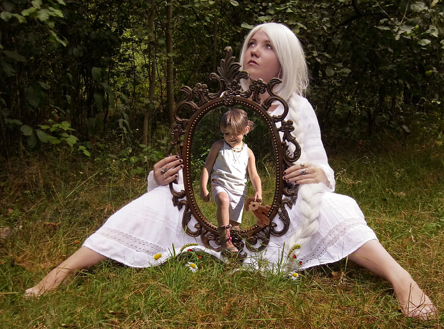 Woman with white wig holding mirror reflecting child in forest