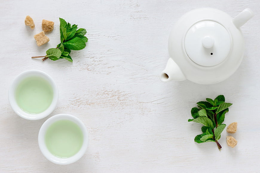 Top view of white teapot, green tea cups, and fresh mint leaves on white wood