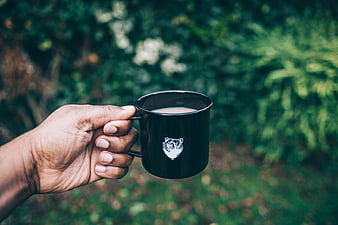 Hand holding black camping mug with coffee outdoors