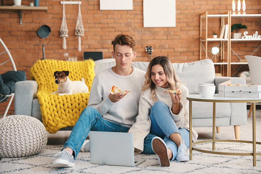 Young couple eating pizza on floor with laptop and dog