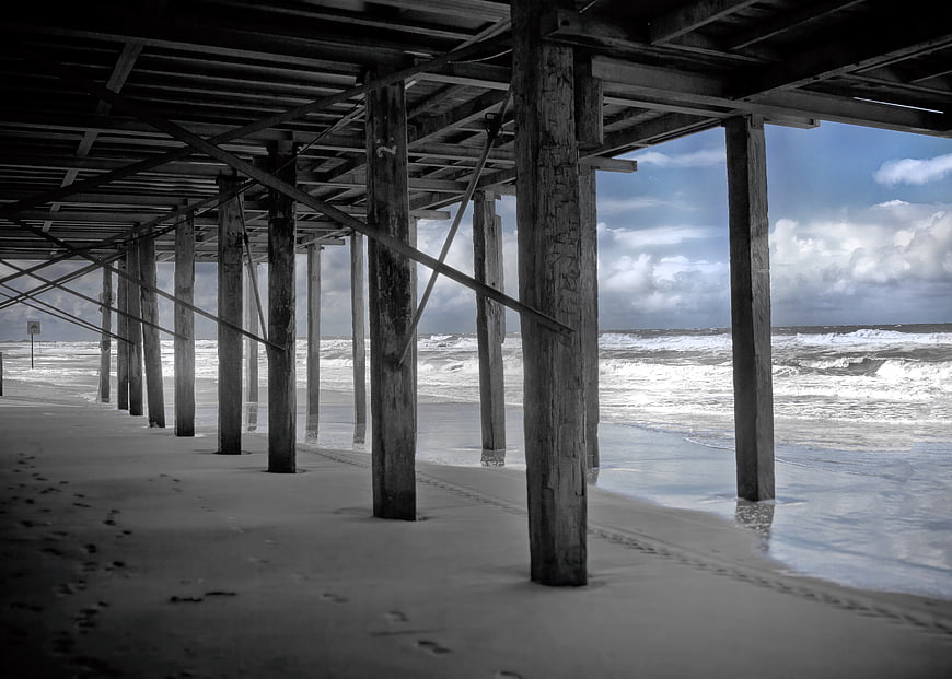 Underneath a weathered wooden pier on a sandy beach with ocean waves