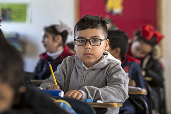 Young boy with glasses holding a pencil in a classroom