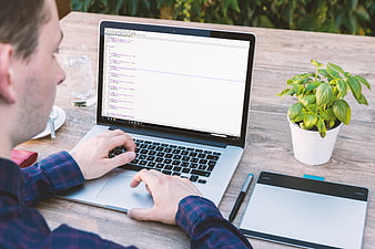 Man coding on laptop at wooden table with tablet and plant