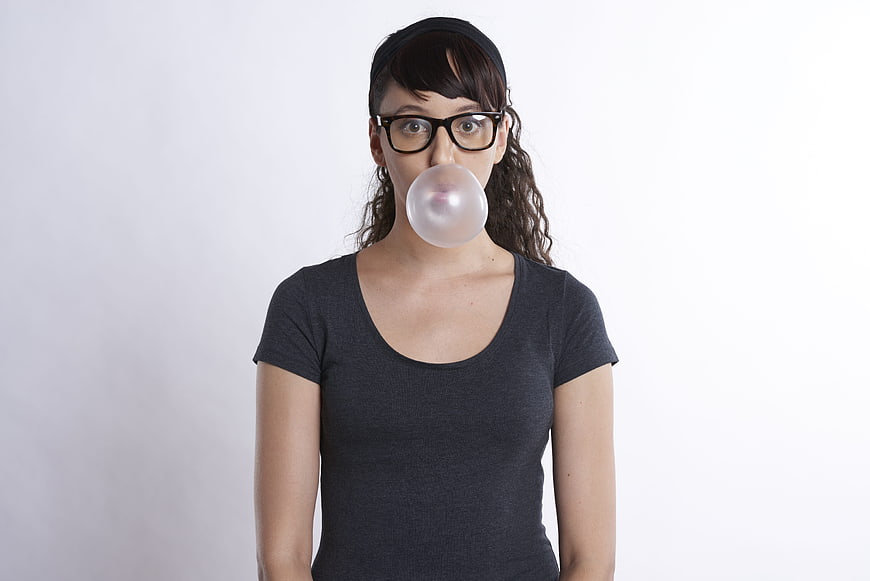 Young woman with glasses blowing a bubblegum bubble against a white background