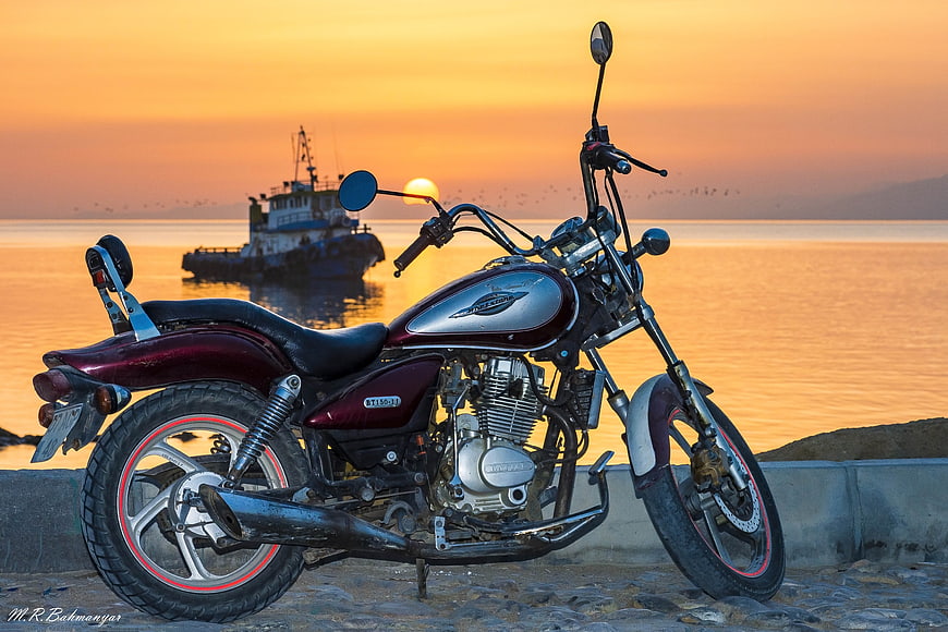 Maroon motorcycle parked by the sea at sunset with fishing boat