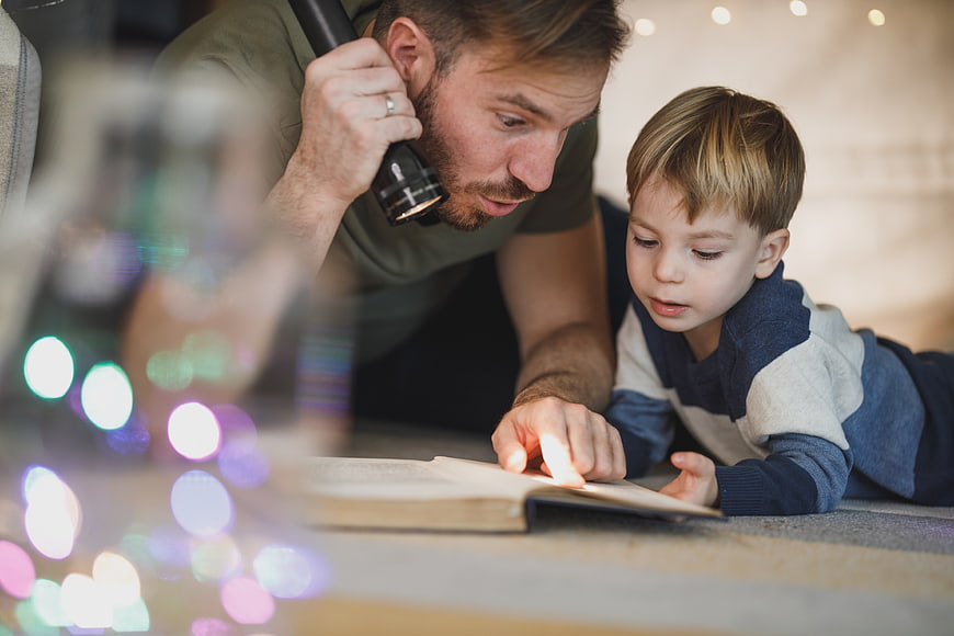 Father and son reading book with flashlight indoors