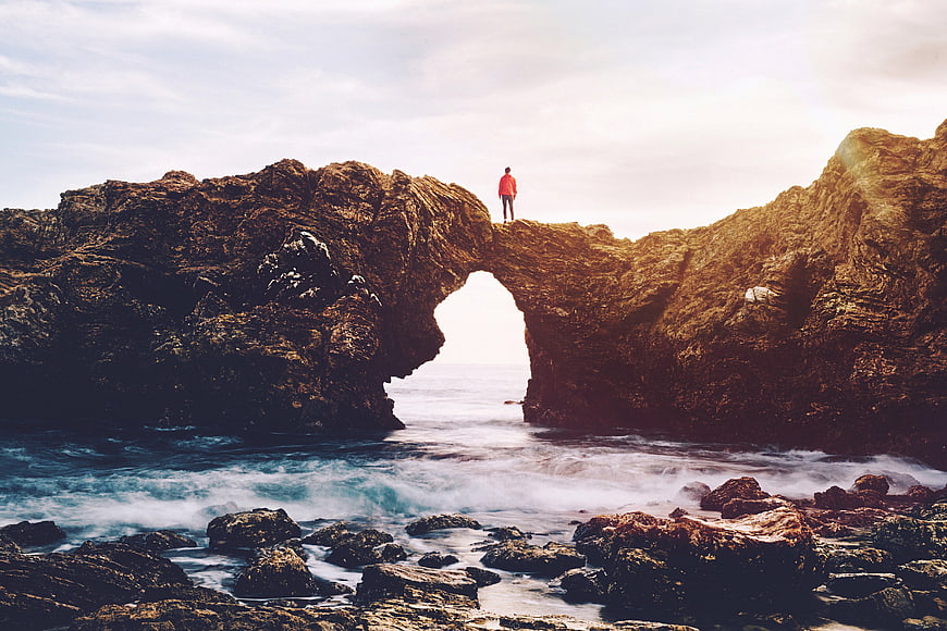 Man standing on coastal rock arch overlooking ocean waves