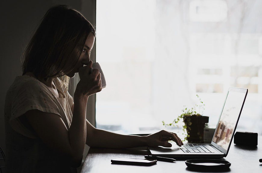 Woman drinking coffee while working on laptop by window