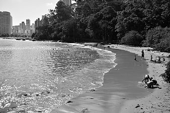 Black and white photo of people relaxing on a sandy beach near city skyline