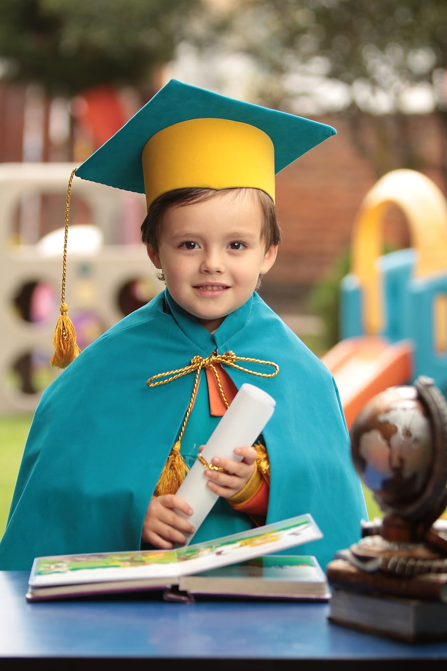 Young child in teal graduation cap and gown holding diploma outdoors