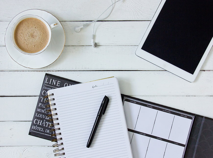 Flat lay of coffee, notebook, tablet, and headphones on white wood