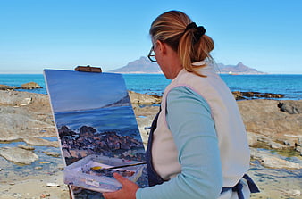 Woman painting coastal landscape on easel at rocky beach
