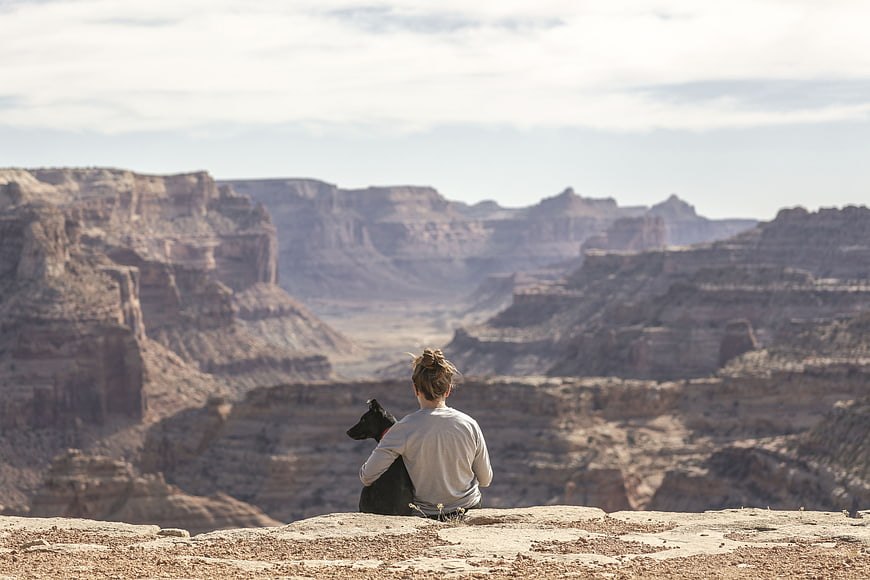 Woman and dog sitting on canyon rim overlooking vast desert landscape