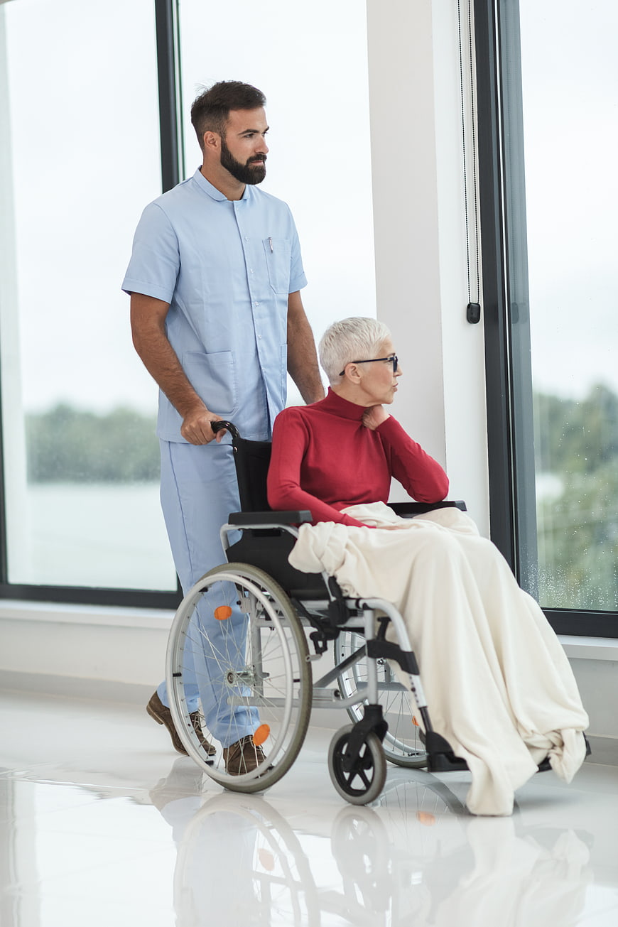 Male nurse pushing elderly woman in wheelchair near window