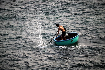 Shirtless man bailing water from small blue boat in choppy sea