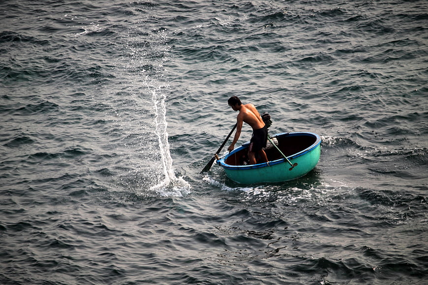 Shirtless man bailing water from small blue boat in choppy sea