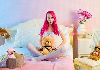 Woman with pink hair sitting on bed with teddy bears