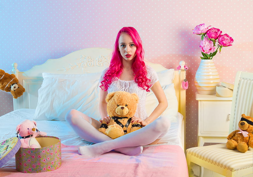 Woman with pink hair sitting on bed with teddy bears