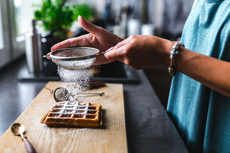 Woman sifting powdered sugar over waffle on wooden board