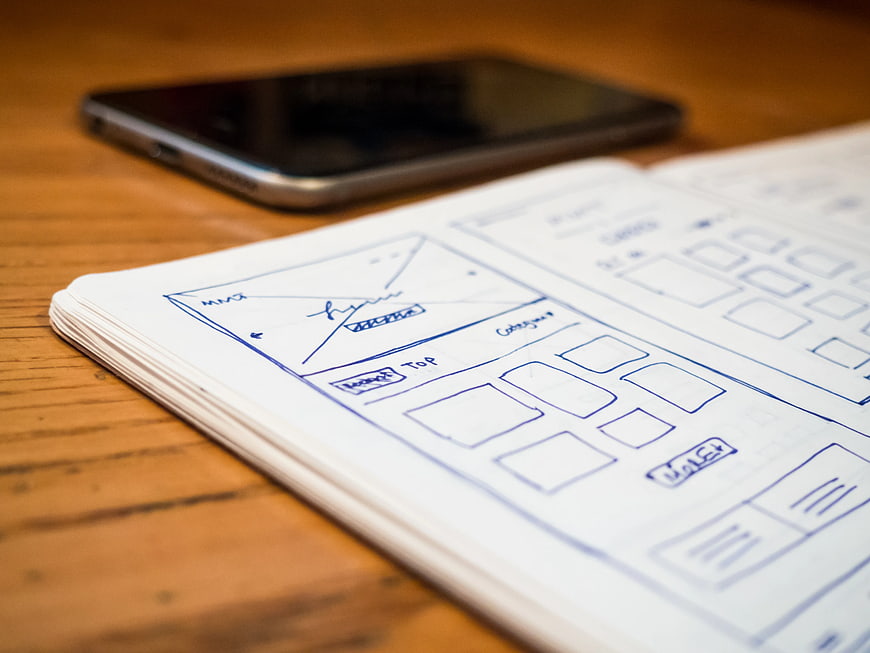 Hand-drawn UI wireframes in sketchbook with smartphone on wooden desk