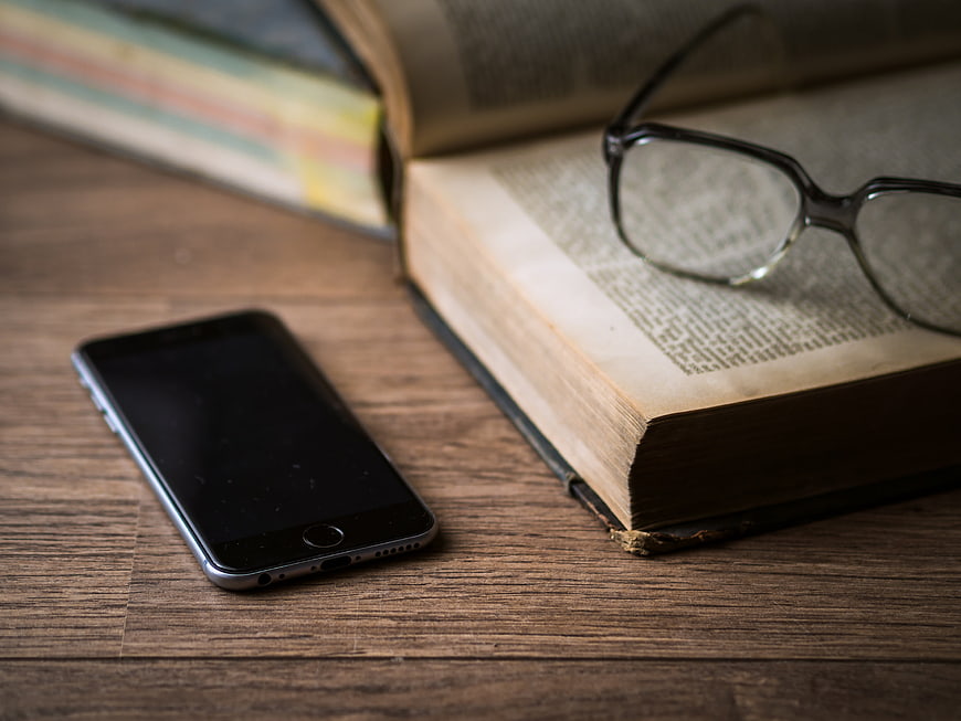 Smartphone and Reading Glasses on Open Book on Wooden Desk