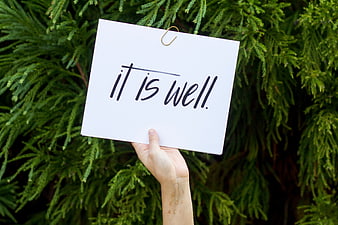 Hand holding sign saying it is well against green foliage