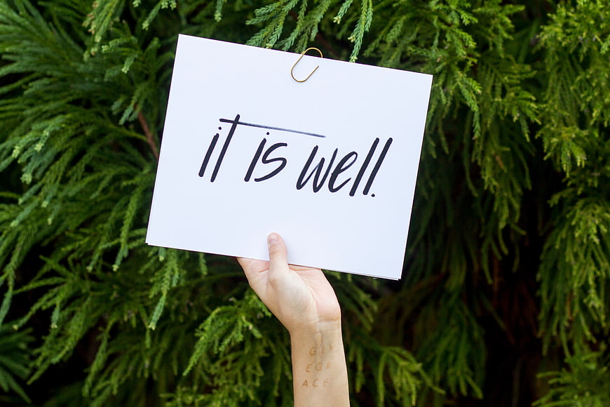 Hand holding sign saying it is well against green foliage
