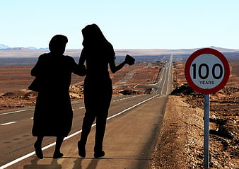 Silhouettes of Two Women Walking on Desert Highway Near 100 Years Sign