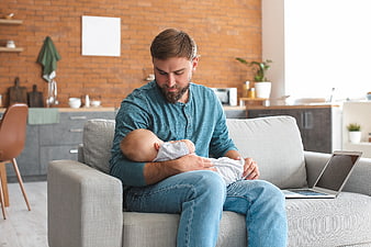 Father holding sleeping baby on couch with laptop