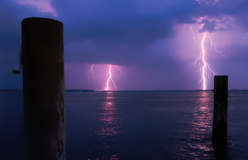 Dramatic purple lightning storm over calm ocean water with pier posts