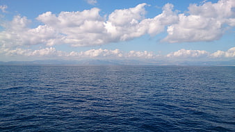 Serene blue ocean horizon with fluffy white clouds and distant mountains