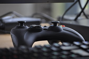 Black Game Controller On Desk With Blurred Keyboard And Monitor