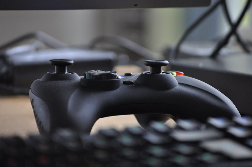 Black Game Controller On Desk With Blurred Keyboard And Monitor