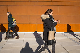 Two women walking past orange wall using smartphone