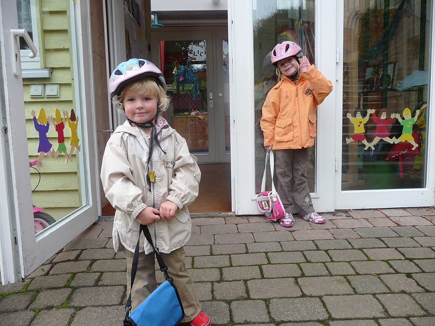 Two young children wearing bike helmets standing outside a kindergarten building