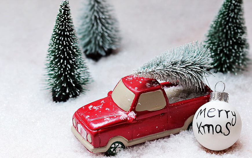 Red toy truck carrying Christmas tree in snow with ornament